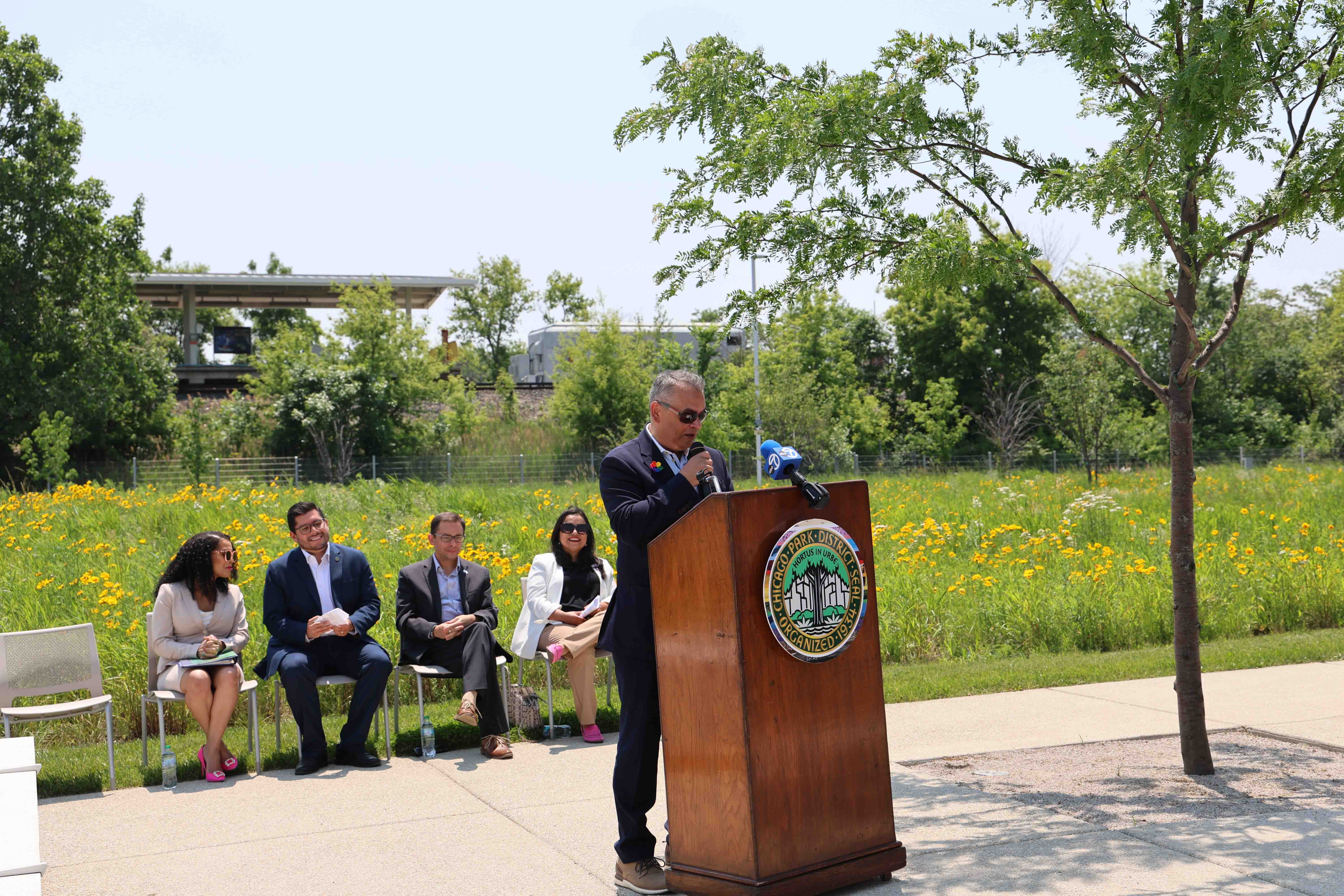 Man speaks at podium with Chicago Park District seal; audience sits nearby. Train platform and wildflowers in background.